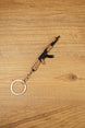 Miniature AK-74 rifle keyring angled on wood background, showing curved magazine, black body and wood-effect rifle details