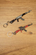 Base black AKM rifle keychain and red modded rifle keyring displayed diagonally on wood with detailed miniature parts visible