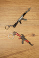 Two AKM-style rifle keyrings with metal chain attachments shown at an angle on wood, ideal for FPS gamers and mil-sim fans