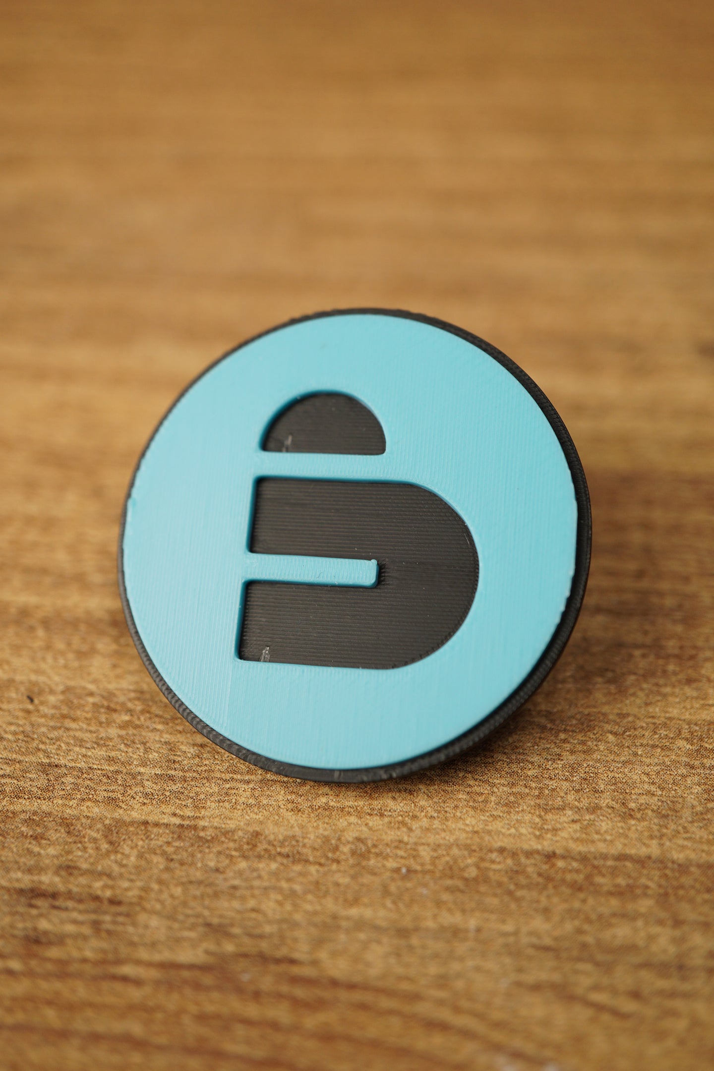 Close-up of a blue and black Arc Raiders inspired round pin with bold cutout-style emblem, displayed on a wood grain tabletop.