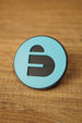 Close-up of a blue and black Arc Raiders inspired round pin with bold cutout-style emblem, displayed on a wood grain tabletop.