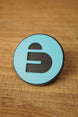 Close-up of a blue and black Arc Raiders inspired round pin with bold cutout-style emblem, displayed on a wood grain tabletop.