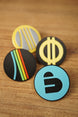 Four Arc Raiders inspired collectible pins arranged on a wooden tabletop, showing colorful circular designs with raised emblem details.