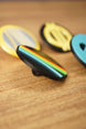 Angled close-up of a black Arc Raiders inspired pin with raised multicolor stripe details, with other round pins softly blurred in the background.