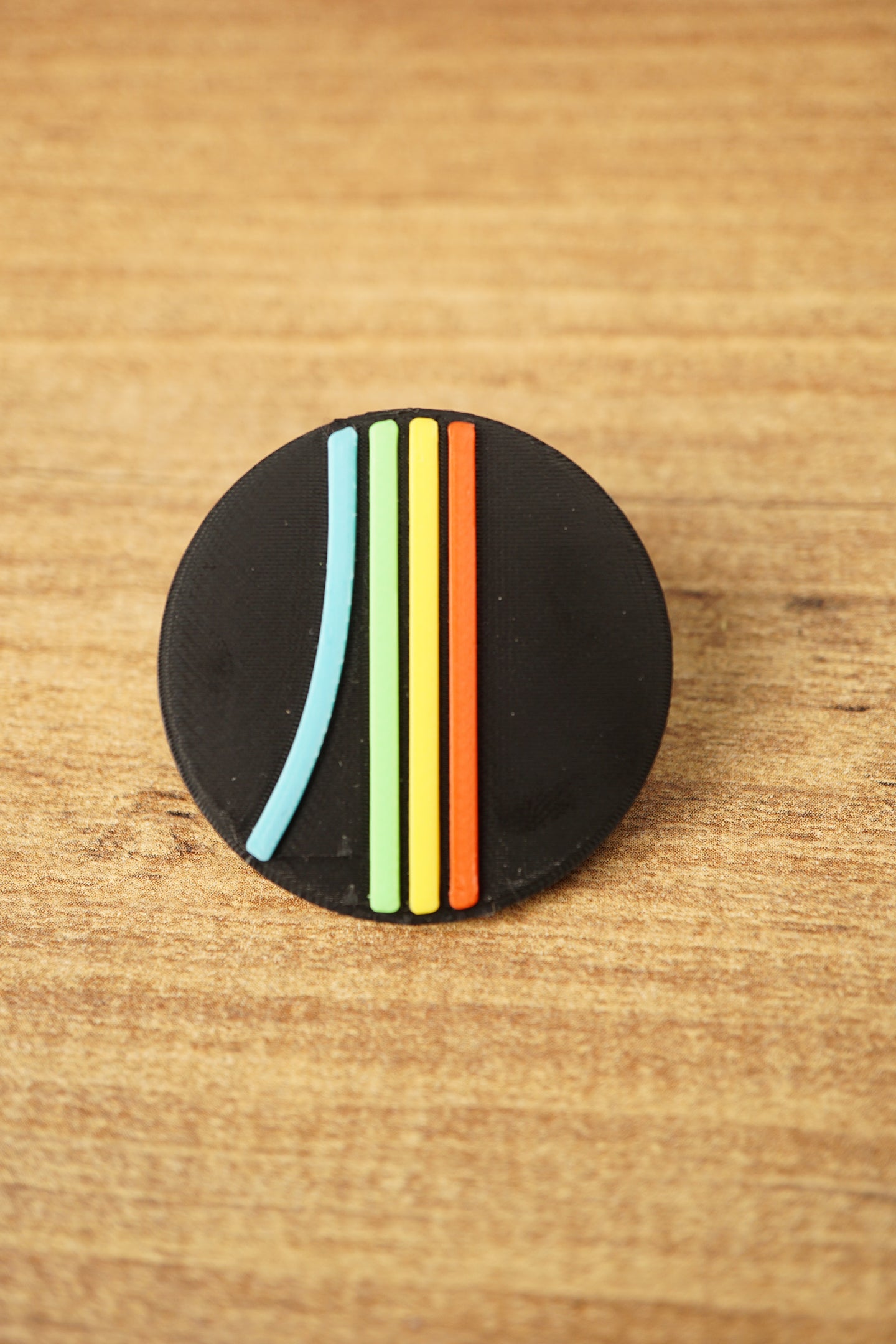 Close-up of a black Arc Raiders inspired round pin with raised blue, green, yellow, and orange stripe details on a wooden background.
