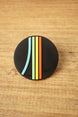 Close-up of a black Arc Raiders inspired round pin with raised blue, green, yellow, and orange stripe details on a wooden background.