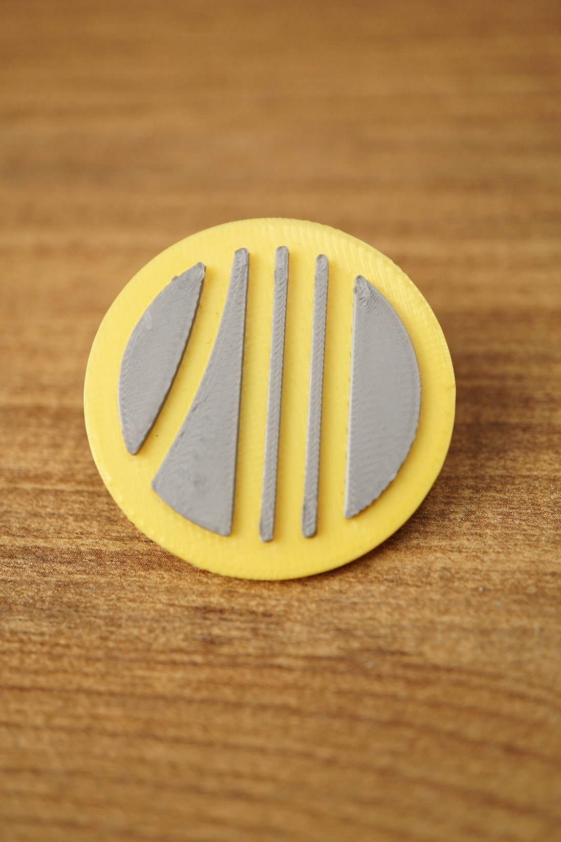 Yellow and grey Arc Raiders inspired circular pin with raised stripe design, photographed upright on a wooden tabletop for product detail.