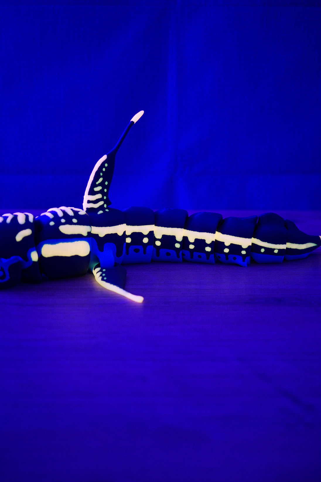 Side view of articulated Marrowbreach creature figure glowing under UV light, showing dark blue body, pale glow markings, fins and segmented tail.