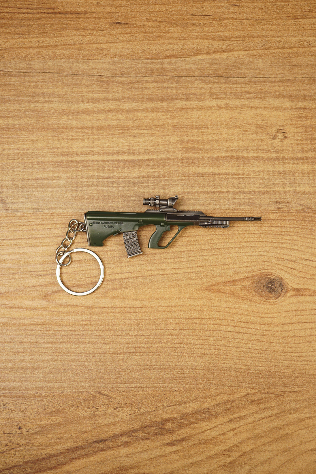AUG-style metal rifle keyring laid flat on a wooden surface, showing the green bullpup body, scope detail and secure keychain ring