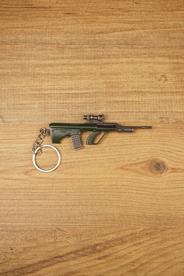 AUG-style metal rifle keyring laid flat on a wooden surface, showing the green bullpup body, scope detail and secure keychain ring