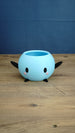 Light blue Stardew Valley Junimo planter pot with black arms, black feet, oval eyes, and pink cheeks on a wooden tabletop.