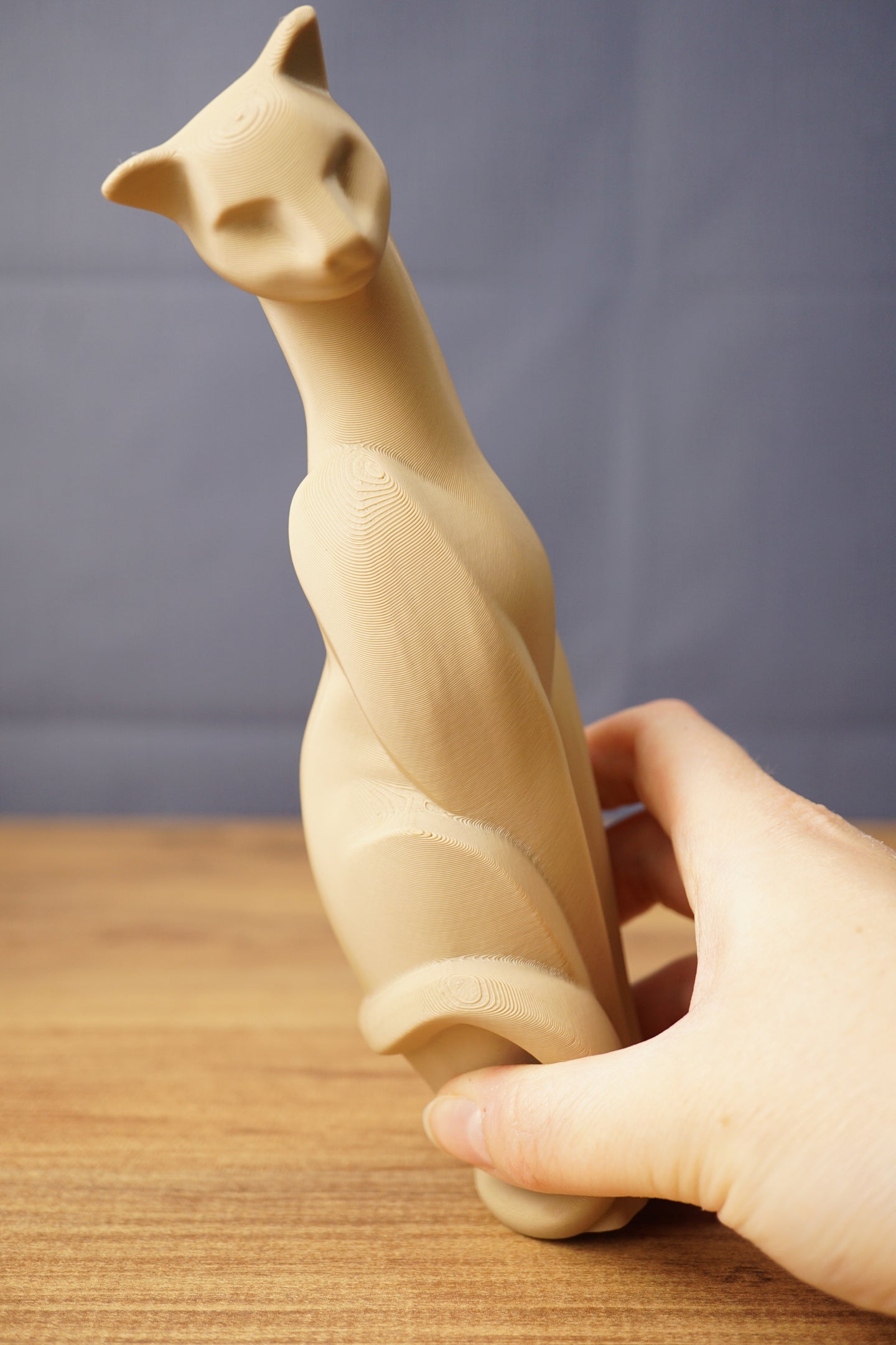 Close-up of a beige 3D printed cat figurine replica held by hand, highlighting the long neck, stylized face, curved body, and print texture.