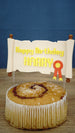 Custom RuneScape quest scroll cake topper on a cinnamon roll cake with yellow birthday text, red badge detail, and clear acrylic dessert sticks.