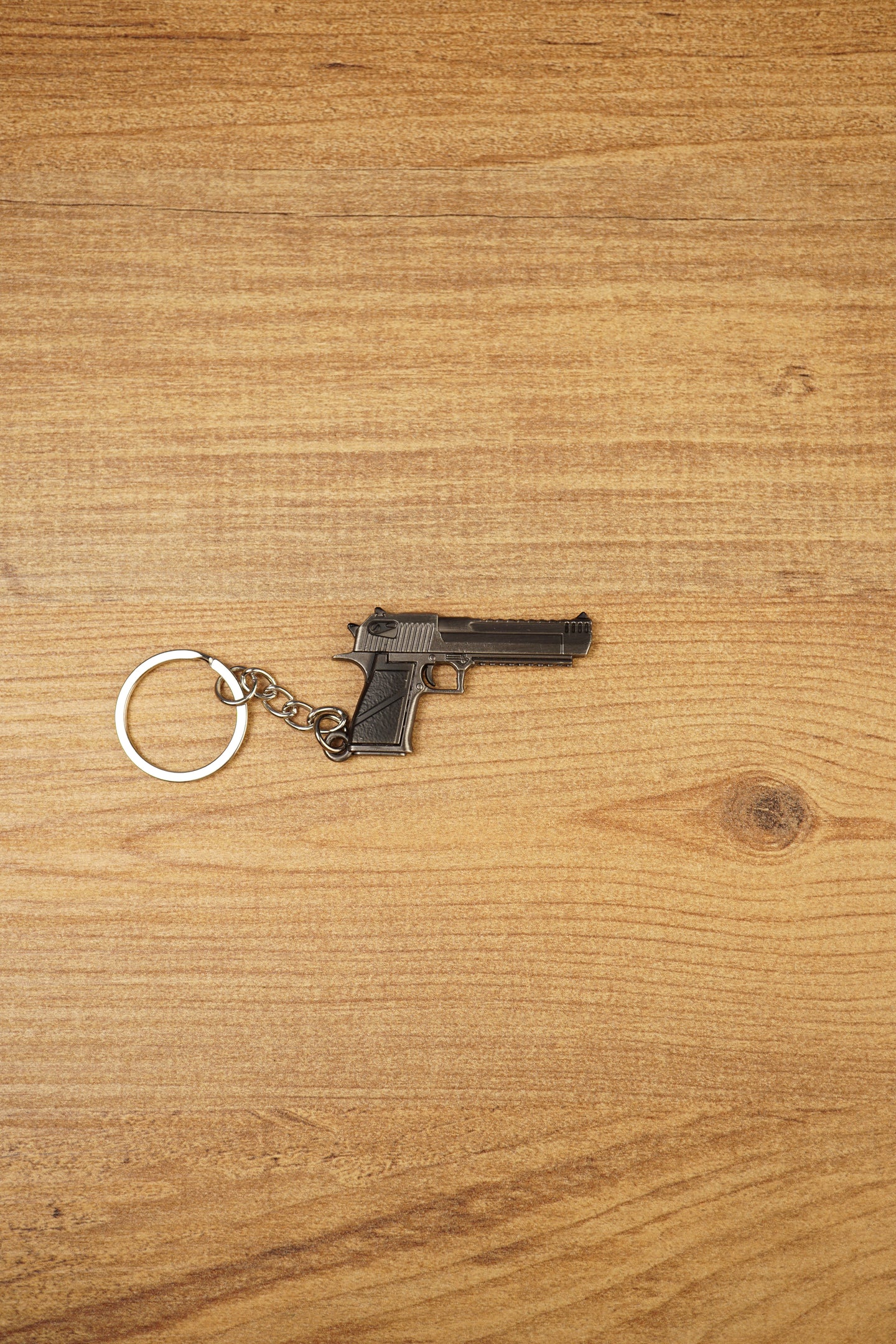 Mini Desert Eagle pistol keychain replica laid flat on a wooden surface, showing the gunmetal finish, black grip and silver split ring attachment