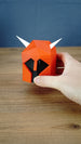 Hand holding the small Dragon Med Helm display replica, showing its compact size, red helmet body, black face and white horns