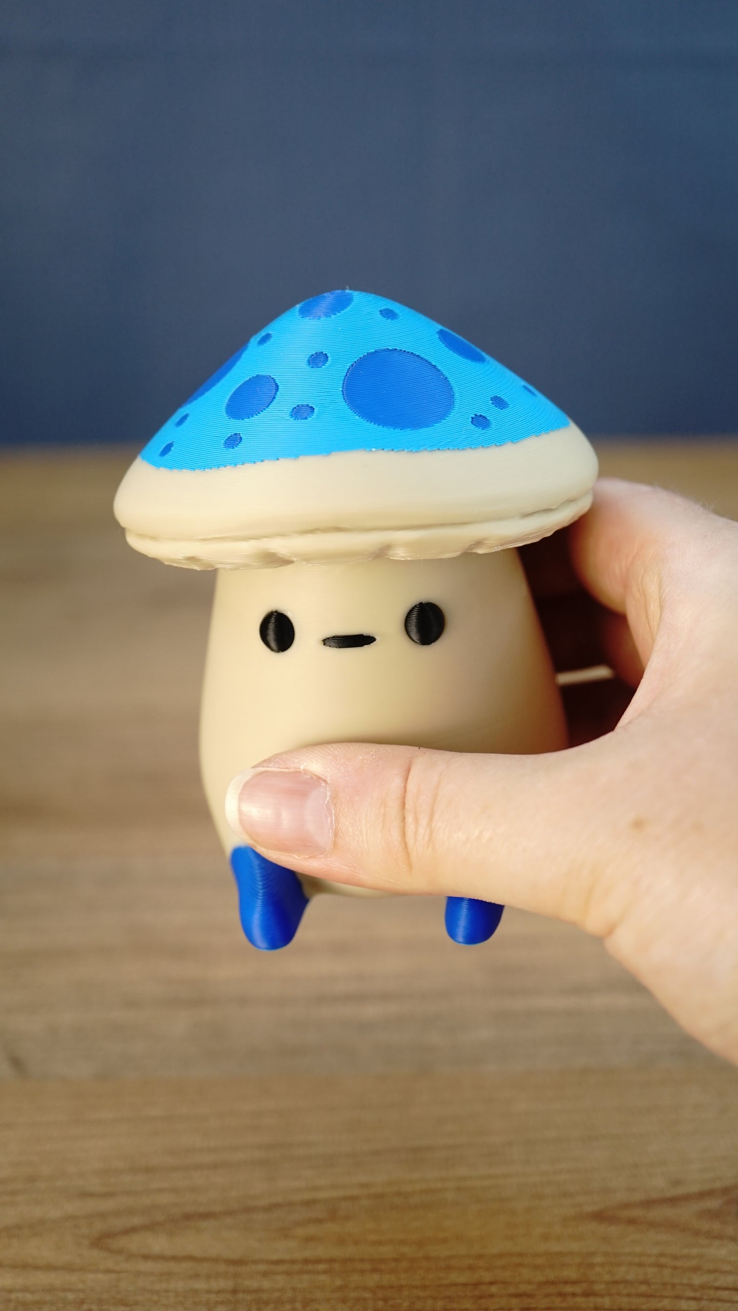 Close-up of an Elderwood Fun Guy figurine being held, showing its blue spotted cap, small black face, cream body, and blue feet.
