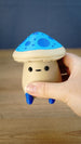 Close-up of an Elderwood Fun Guy figurine being held, showing its blue spotted cap, small black face, cream body, and blue feet.