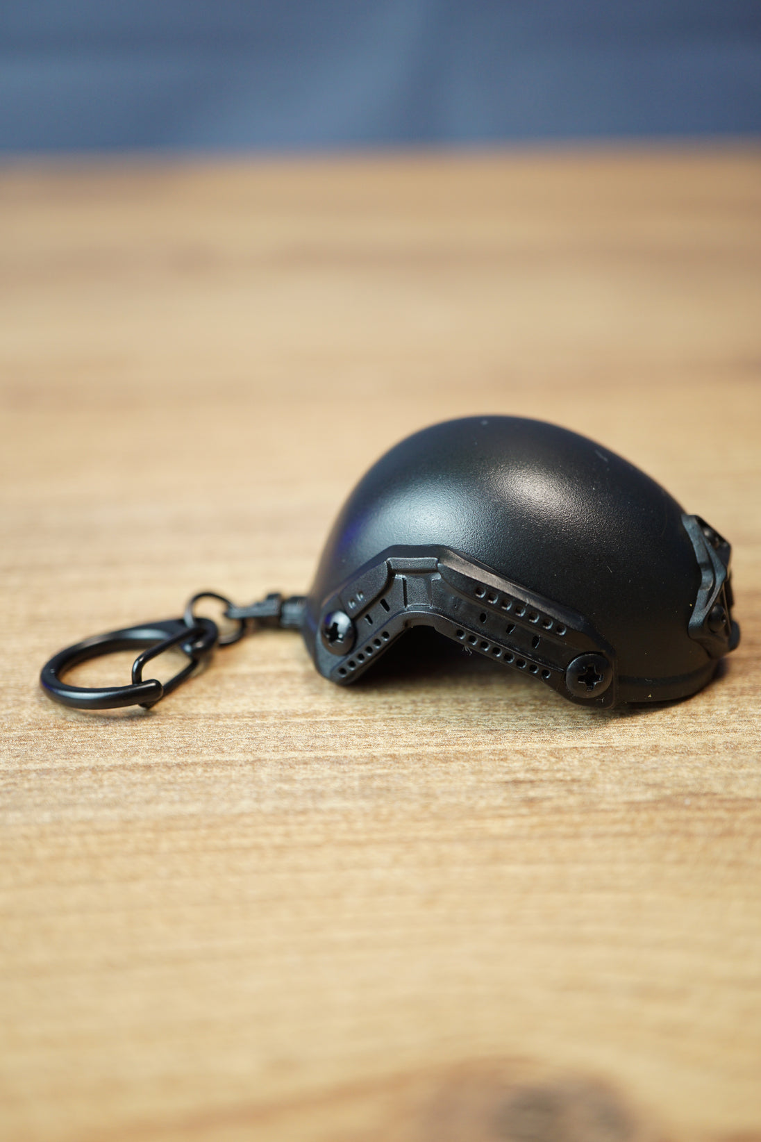 Side view of a FAST MT helmet bottle opener keychain featuring a black helmet shell, rail detail, attached keyring, and wooden tabletop background.