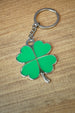 Lucky green four leaf clover keychain with silver outline and attached keyring, photographed from above on a wood grain tabletop.