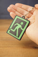 Hand holding a green and black extraction sign keyring, showing the raised 3D printed running figure design and metal keychain.