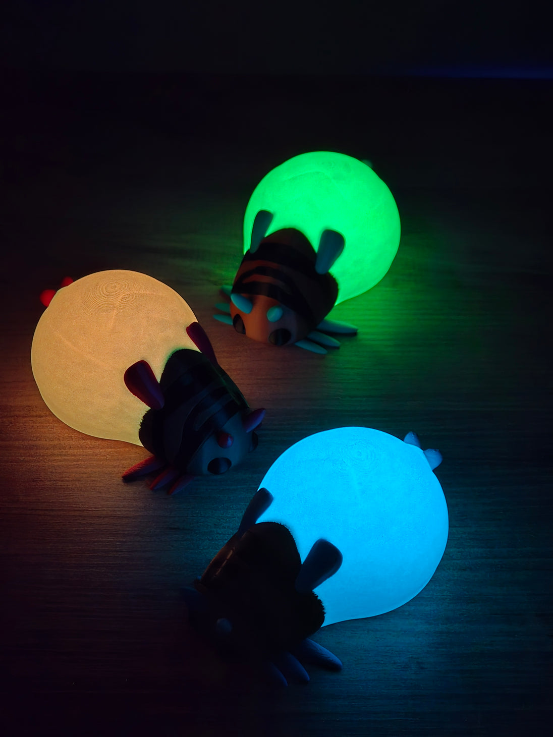 Glow-in-the-dark Palia-inspired lantern bug plush figures glowing in blue, green, and yellow in a dark room.