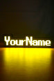 Illuminated YourName LED name sign glowing bright yellow in a dark room with a nostalgic RuneScape-style font design.