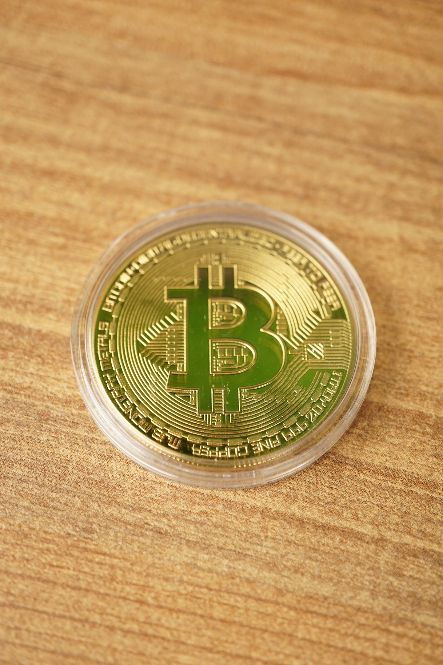 Close-up of a gold Bitcoin replica coin in a clear capsule on a wooden surface, featuring the raised Bitcoin symbol.