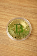 Close-up of a gold Bitcoin replica coin in a clear capsule on a wooden surface, featuring the raised Bitcoin symbol.