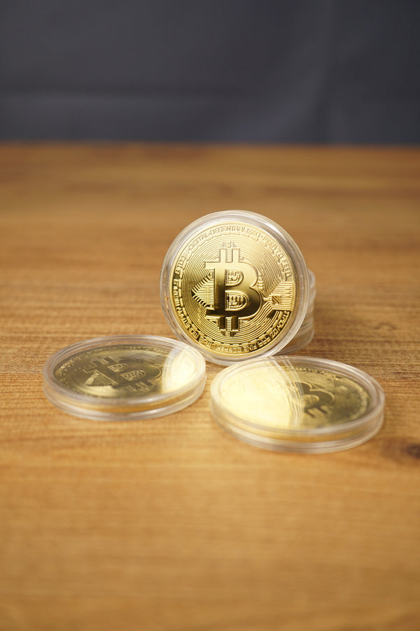 Gold Bitcoin replica coins in clear cases arranged on a wooden tabletop, with one coin upright and two lying flat in front.