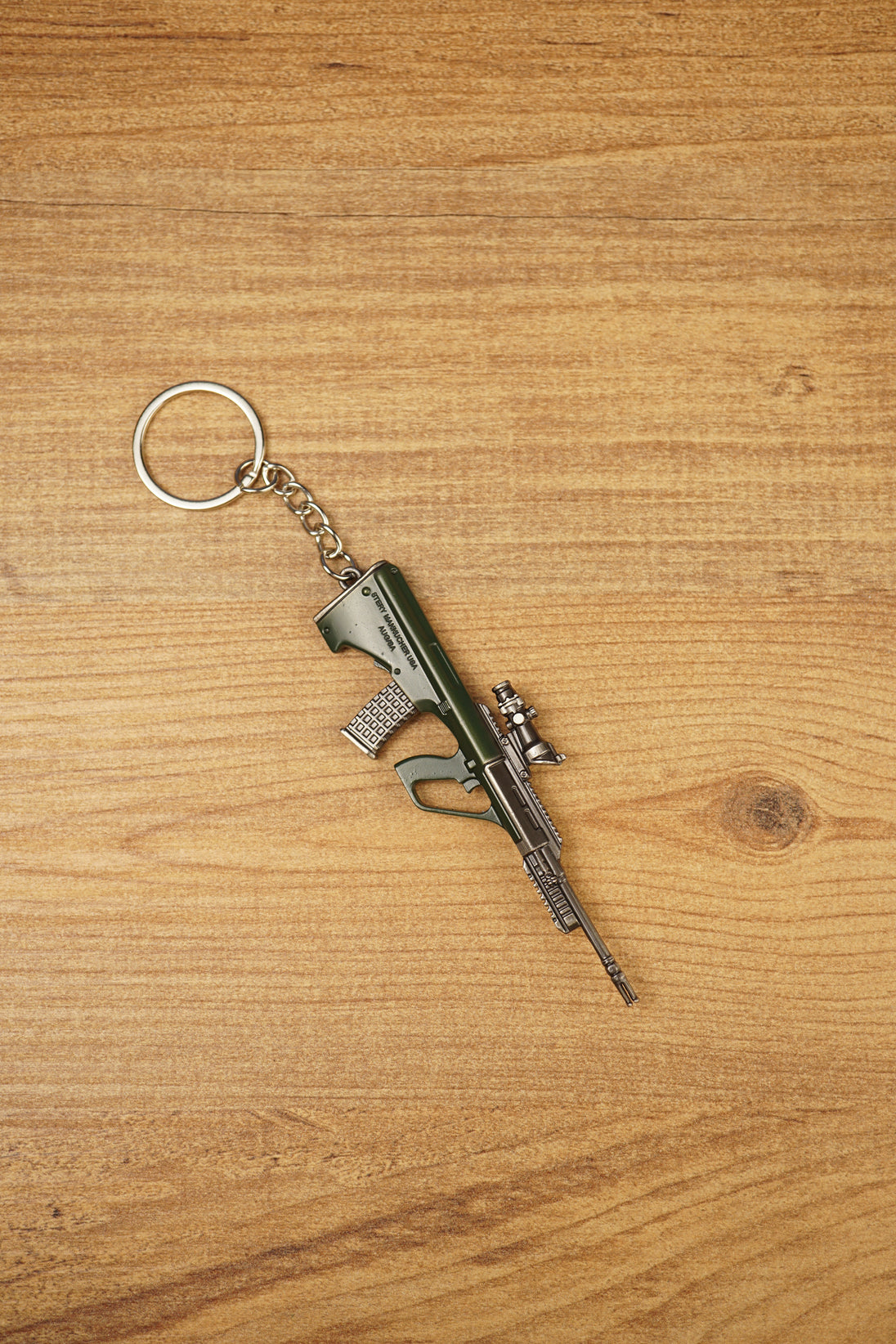Close-up angled view of AUG-style bullpup gun keychain with detailed magazine, optic and metal keyring chain on wood surface
