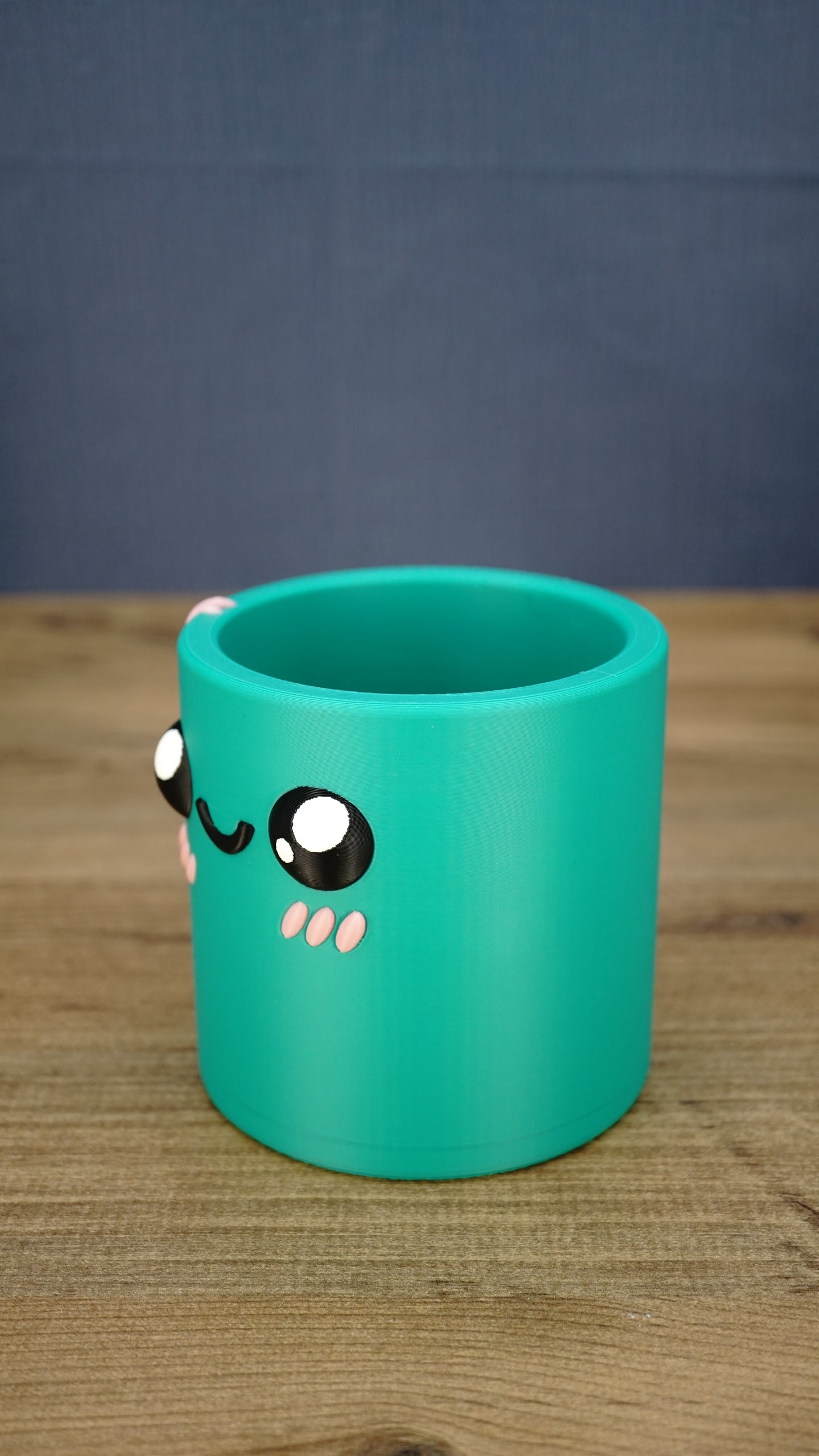 Left-side view of the green Cyl pen pot showing the curved cylindrical body, raised character eyes, pink cheek details and open storage compartment.