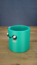 Left-side view of the green Cyl pen pot showing the curved cylindrical body, raised character eyes, pink cheek details and open storage compartment.