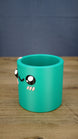 Left-side view of the green Cyl pen pot showing the curved cylindrical body, raised character eyes, pink cheek details and open storage compartment.