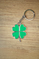 Green four leaf clover keychain with silver metal trim and chain, displayed flat on a wooden surface for a lucky charm accessory photo.
