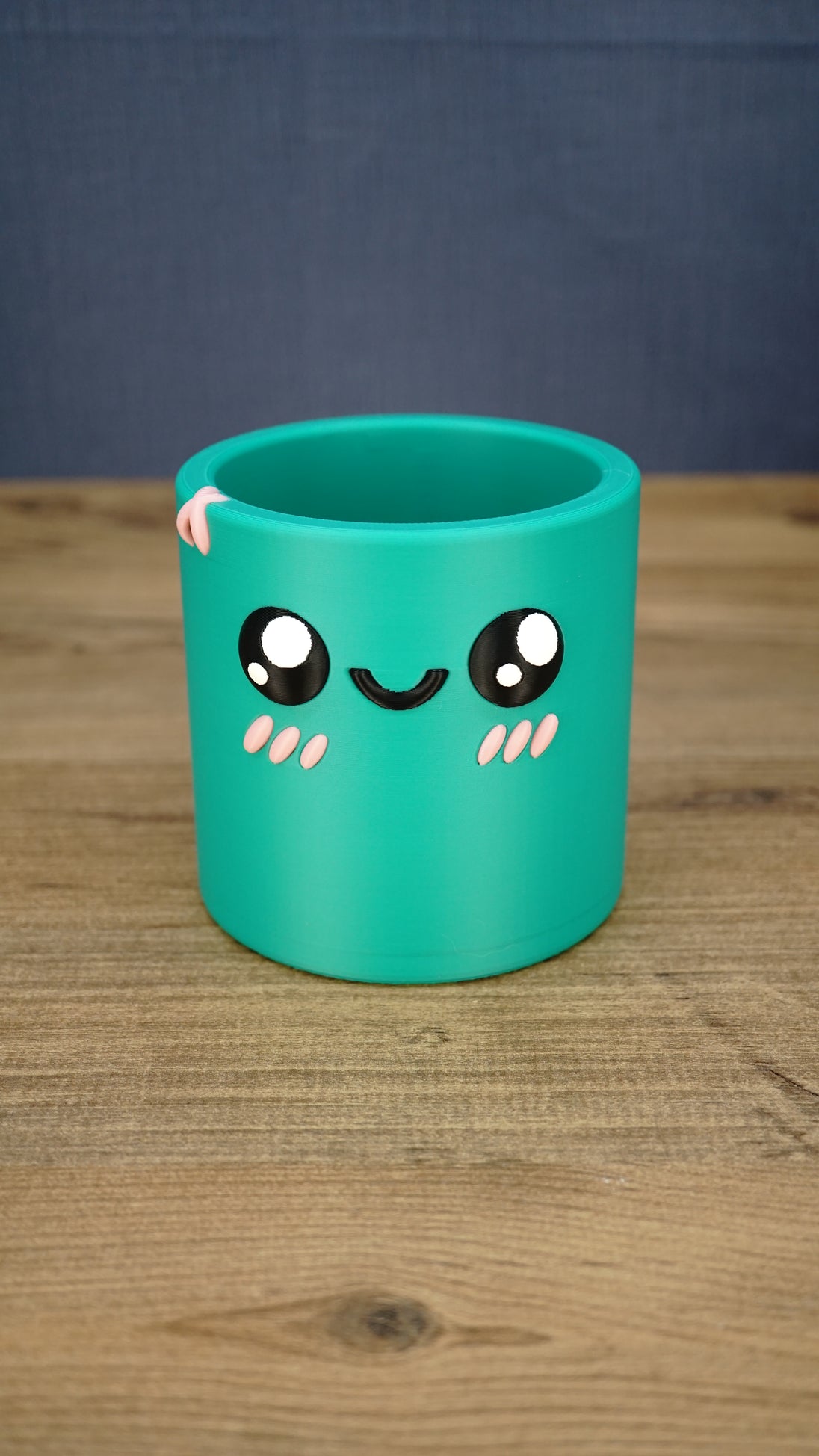 Green Palia-inspired Cyl pen pot shown empty from the front, featuring large black eyes, a smiling face, pink cheeks and decorative rim detail