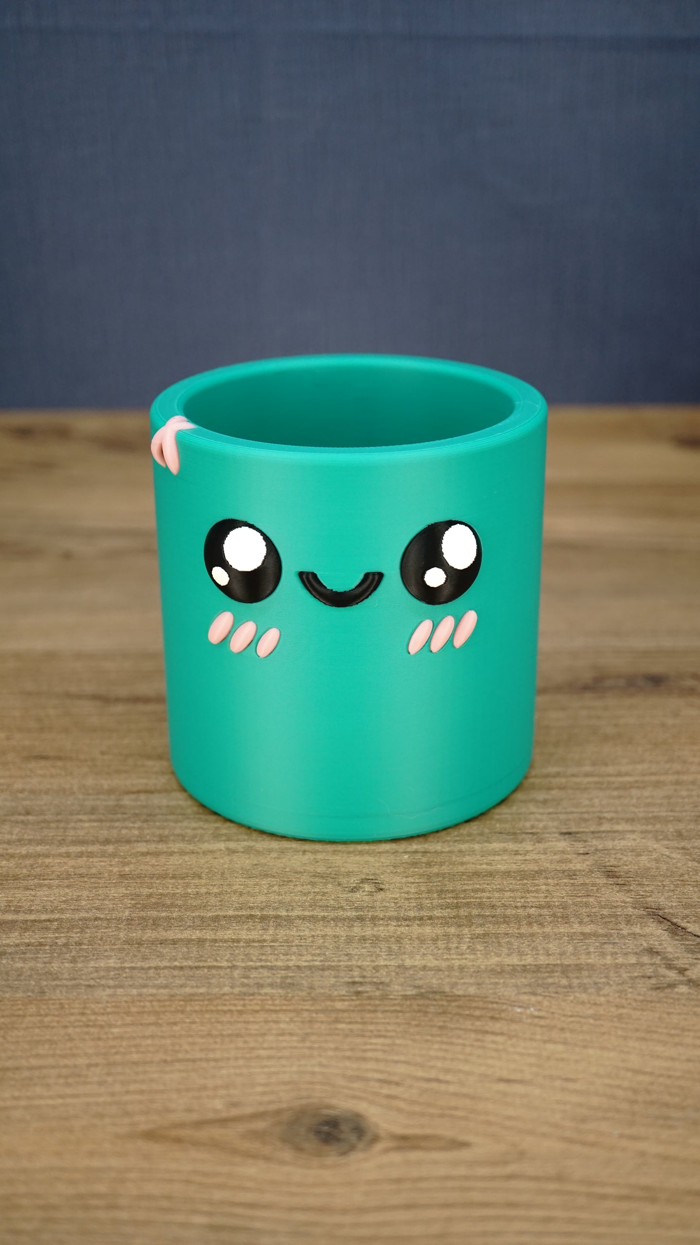 Green Palia-inspired Cyl pen pot shown empty from the front, featuring large black eyes, a smiling face, pink cheeks and decorative rim detail
