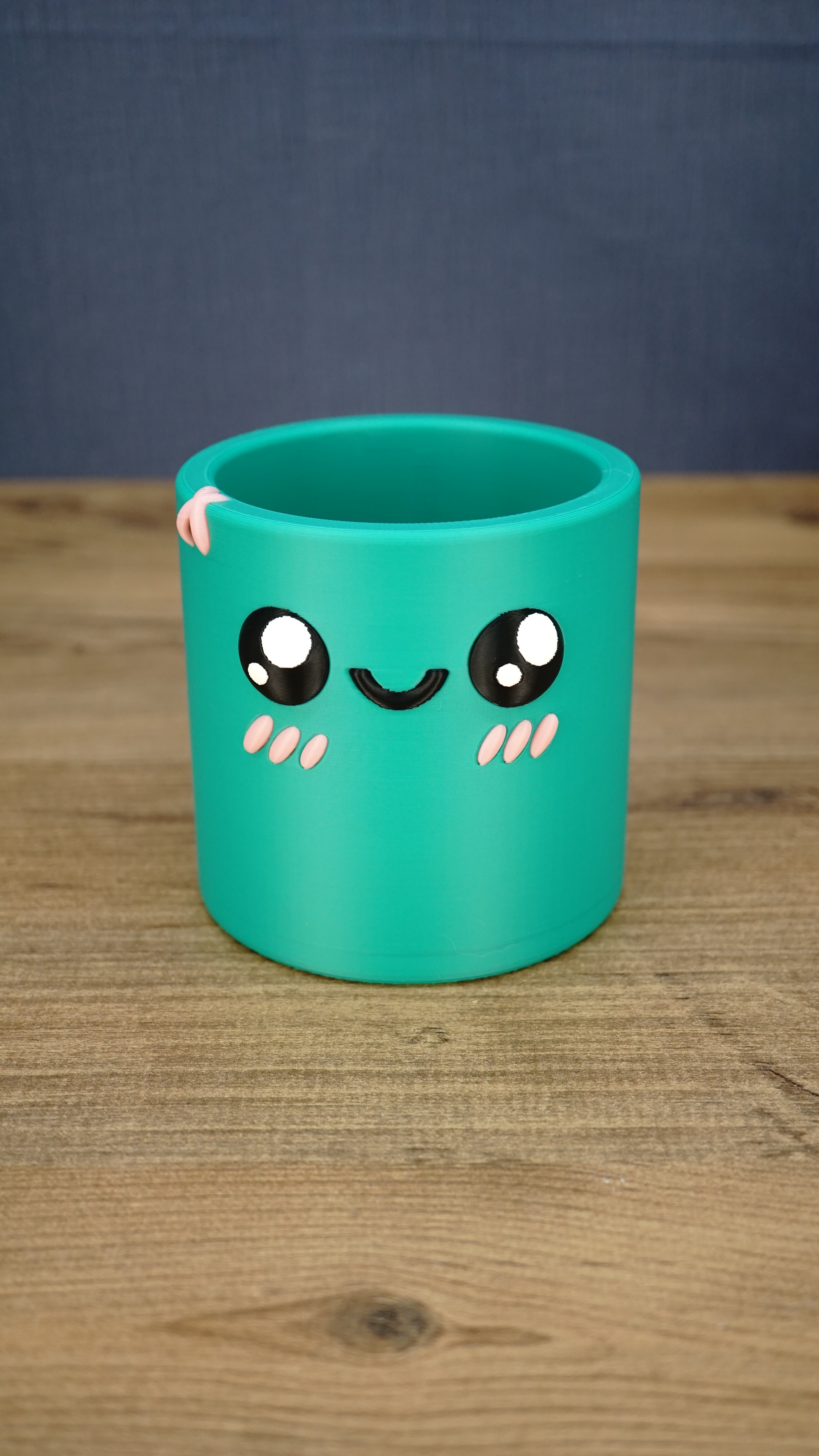 Green Palia-inspired Cyl pen pot shown empty from the front, featuring large black eyes, a smiling face, pink cheeks and decorative rim detail