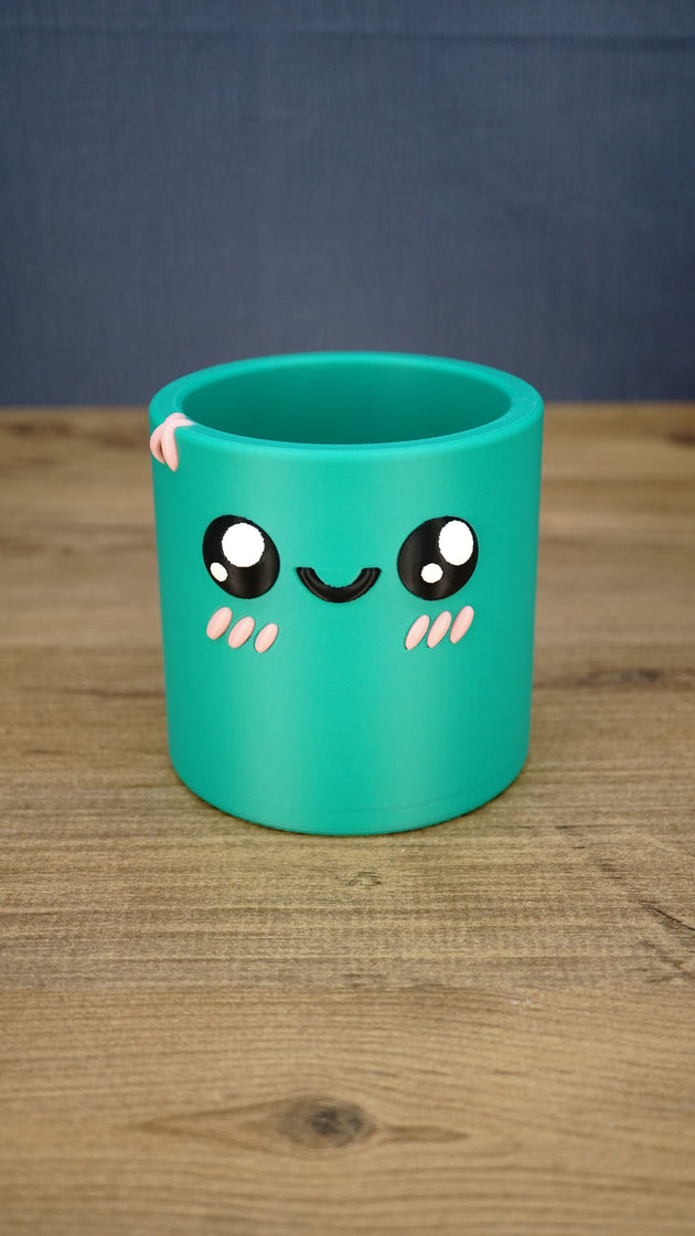 Green Palia-inspired Cyl pen pot shown empty from the front, featuring large black eyes, a smiling face, pink cheeks and decorative rim detail