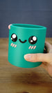 Green Palia-inspired Cyl desk organiser held in one hand to show its size, cylindrical shape, smiling character face and raised decorative details.
