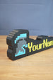 Angled view of a personalized group ironman desk nameplate with a blue and grey helmet icon, yellow RuneScape-style text, and black casing.