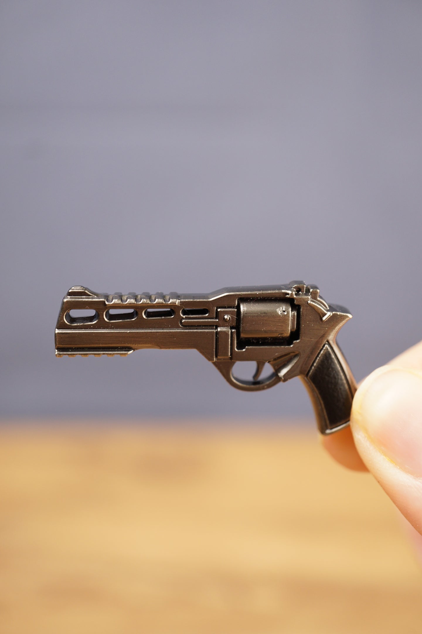 Close-up of compact gunmetal revolver keyring held between fingers, showing the detailed cylinder, barrel, trigger, and black grip