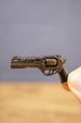 Close-up of compact gunmetal revolver keyring held between fingers, showing the detailed cylinder, barrel, trigger, and black grip
