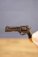 Close-up of compact gunmetal revolver keyring held between fingers, showing the detailed cylinder, barrel, trigger, and black grip