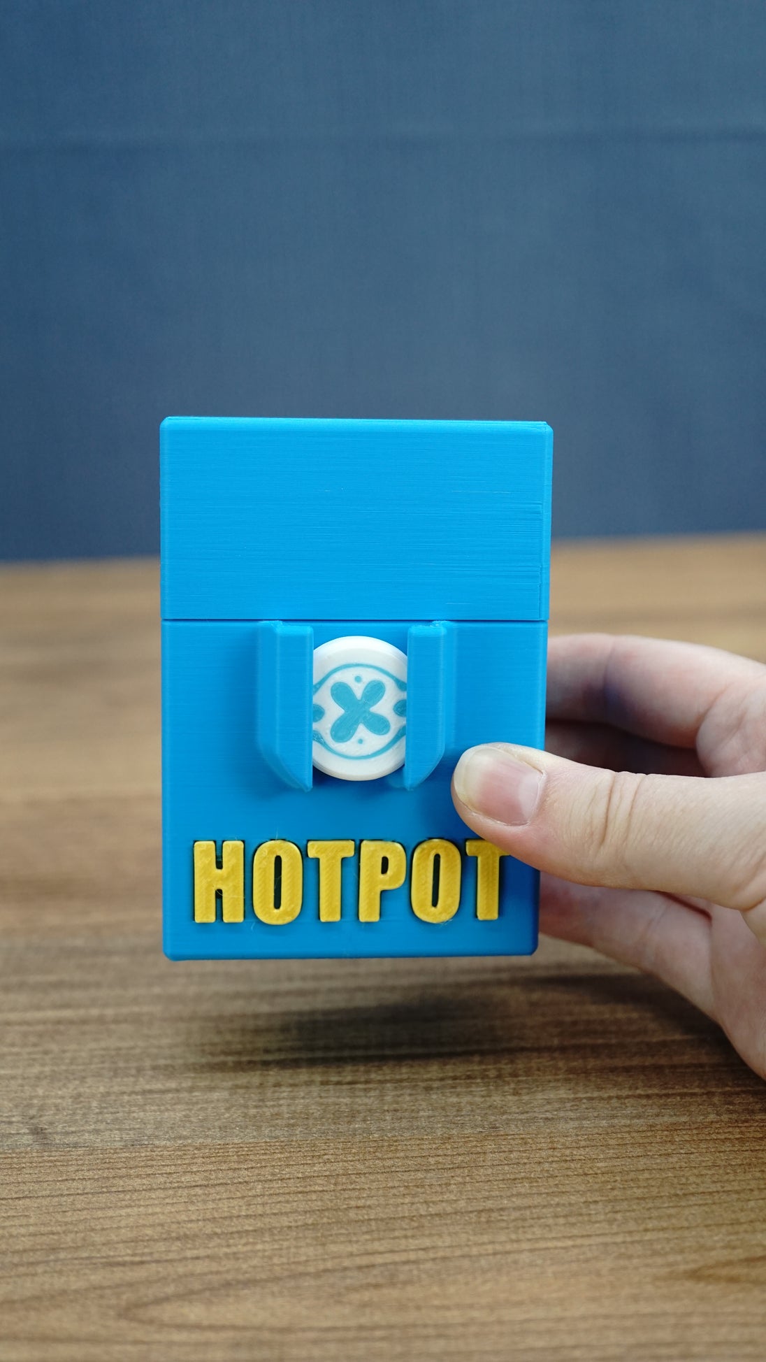 Hand holding a closed blue Palia Hotpot storage case with gold lettering and a white pass token on the front.