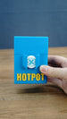 Hand holding a closed blue Palia Hotpot storage case with gold lettering and a white pass token on the front.