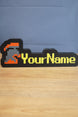 Custom hardcore group ironman nameplate with a red and grey helmet icon, yellow RuneScape-style lettering, and black border on a tabletop.