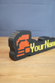 Angled view of a hardcore ironman desk nameplate featuring a red helmet icon, yellow RuneScape-style text, and black outer frame.