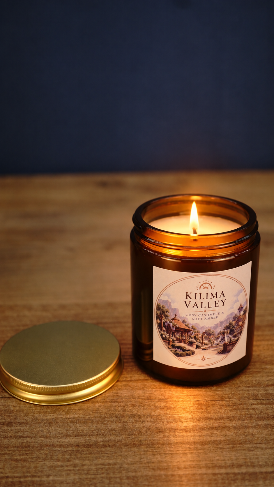 Kilima Valley scented soy wax candle burning in an amber glass jar with its gold lid beside it, featuring cosy cashmere and soft amber fragrance.