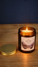 Kilima Valley scented soy wax candle burning in an amber glass jar with its gold lid beside it, featuring cosy cashmere and soft amber fragrance.