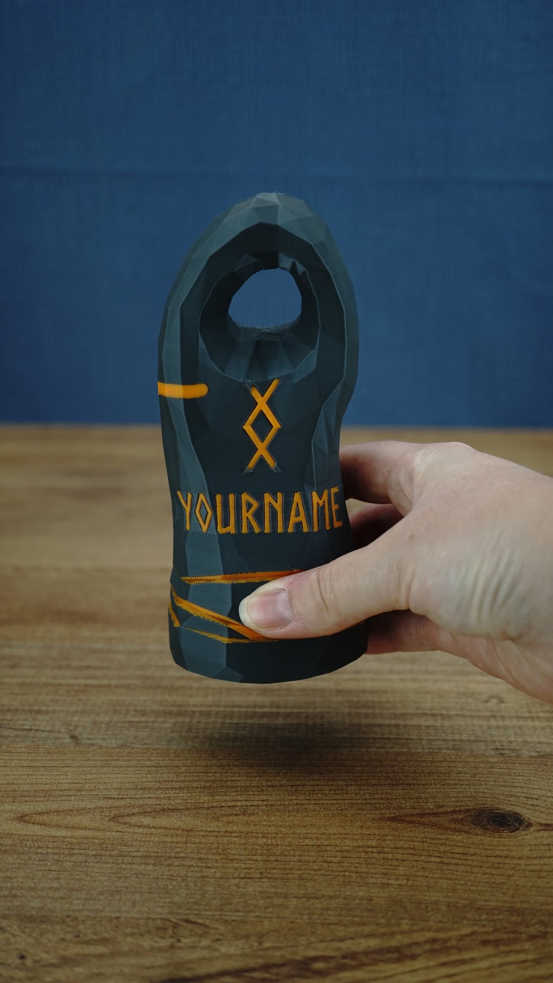 Hand holding a large personalized Valheim-inspired tombstone, showing the orange rune design, YOURNAME text, and low-poly stone texture.
